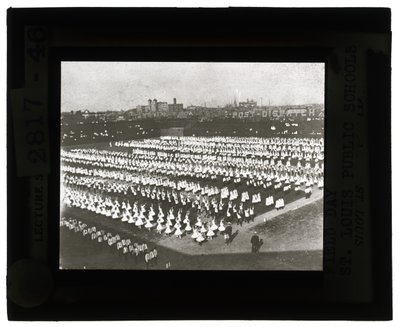 Horizontale, zwart-wit foto van honderden schoolkinderen in formatie voor Field Day parade en spektakel. Alle meisjes in witte jurken en de jongens in witte shirts en stropdassen met donkere broeken door Unbekannt Unbekannt